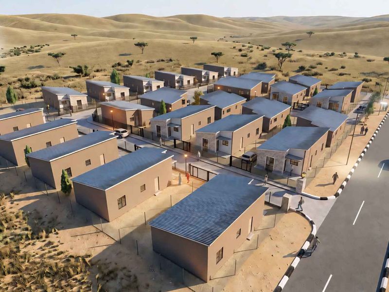 An aerial view of a modern townhouse development featuring multiple residential units, paved access roads, and perimeter security fencing in a desert landscape.