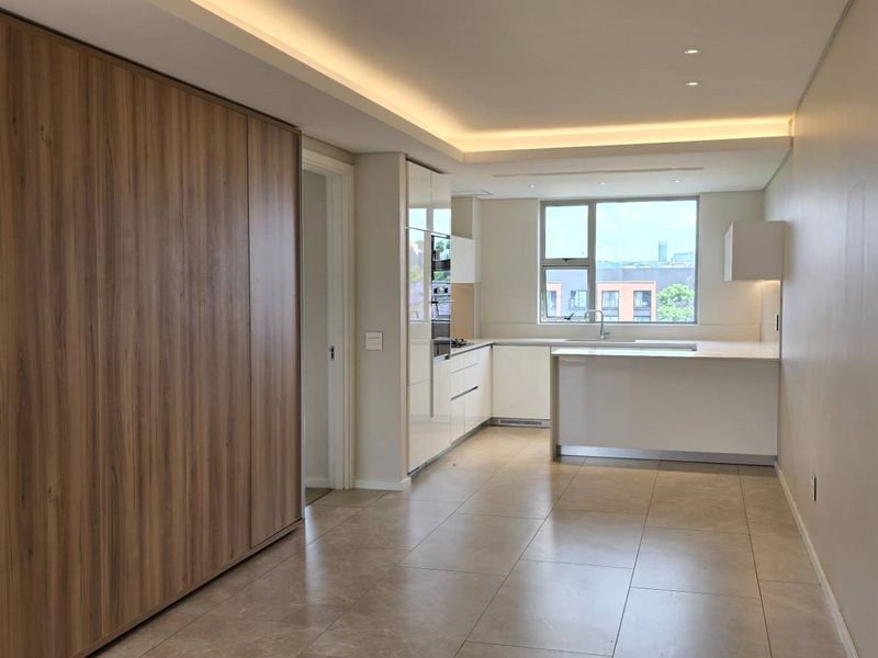 Modern open-plan living area featuring a sleek kitchen with integrated appliances, stone countertops, and recessed ceiling lighting, finished with neutral tiled flooring.