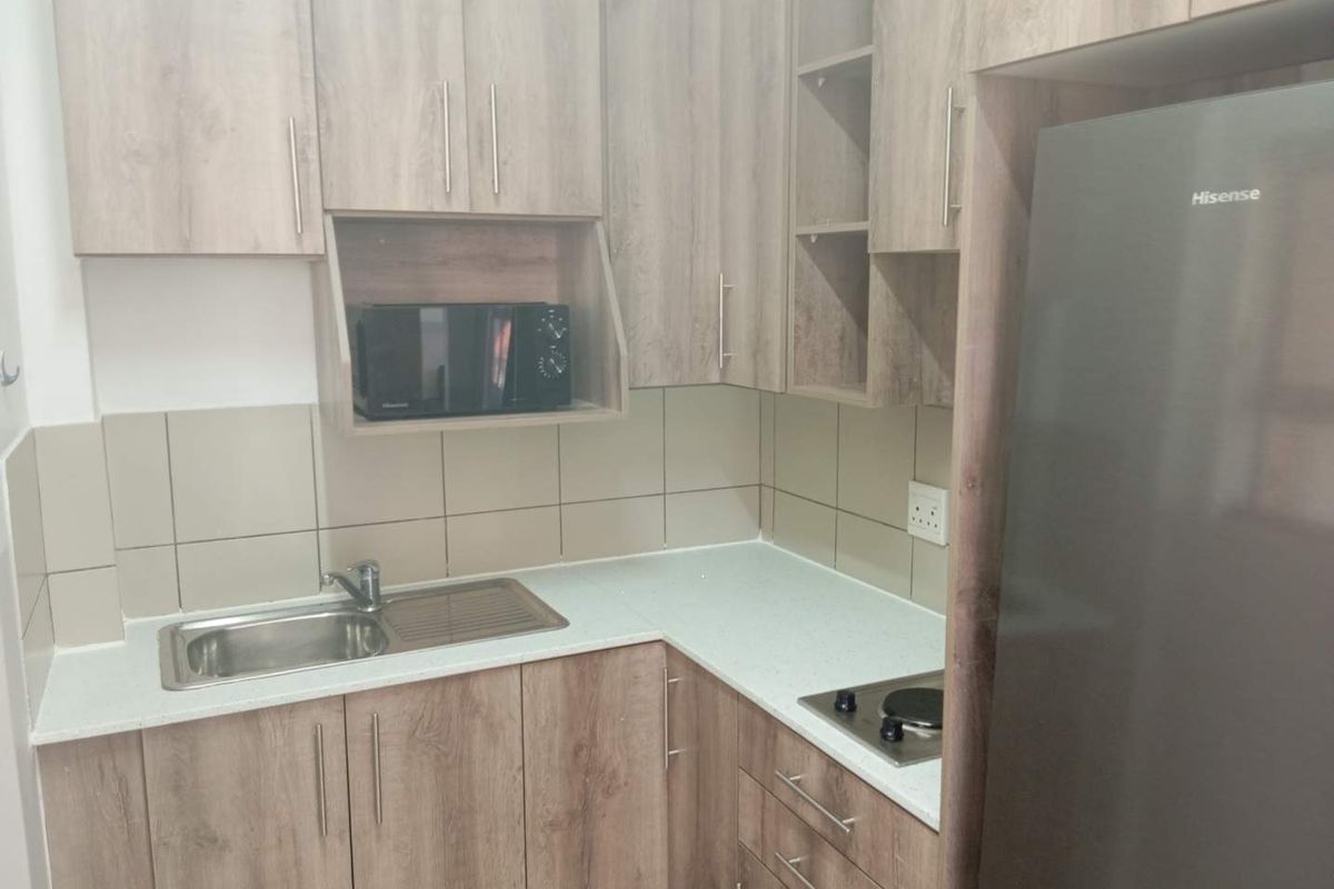 A compact, modern kitchen featuring wood-grain laminate cabinetry, a stainless steel sink, built-in hob, and neutral tiled splashback.