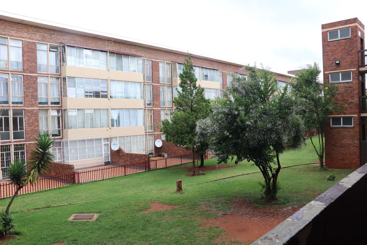 View of the apartment complex exterior featuring brick facade, large windows, and a shared green garden space with mature trees.