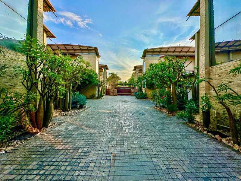 A well-maintained townhouse complex entrance featuring a cobblestone driveway, stone-clad facades, and mature landscaping leading to a secure gated entry.