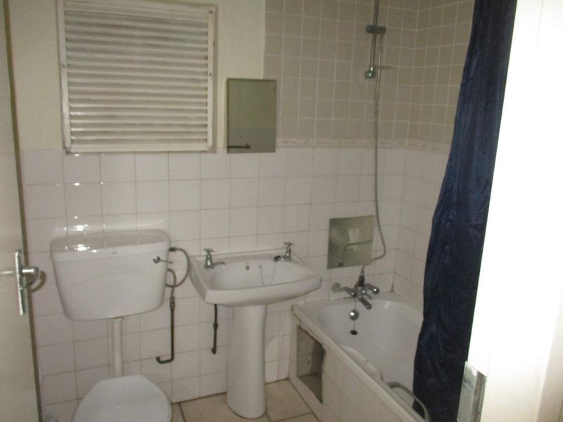 A dated bathroom featuring a pedestal basin, toilet, and bathtub with shower attachment, finished with white wall tiles and a louvered window.
