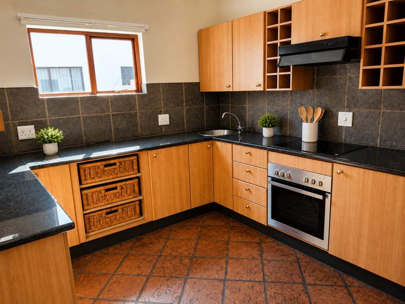 A functional kitchen featuring light wood cabinetry, dark granite countertops, built-in oven with hob, and terracotta floor tiles.
