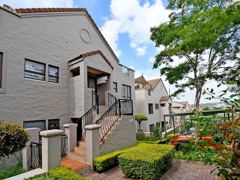 Exterior view of a multi-level apartment complex featuring textured grey facades, terracotta roof tiles, wrought iron railings, and well-maintained landscaped garden areas.