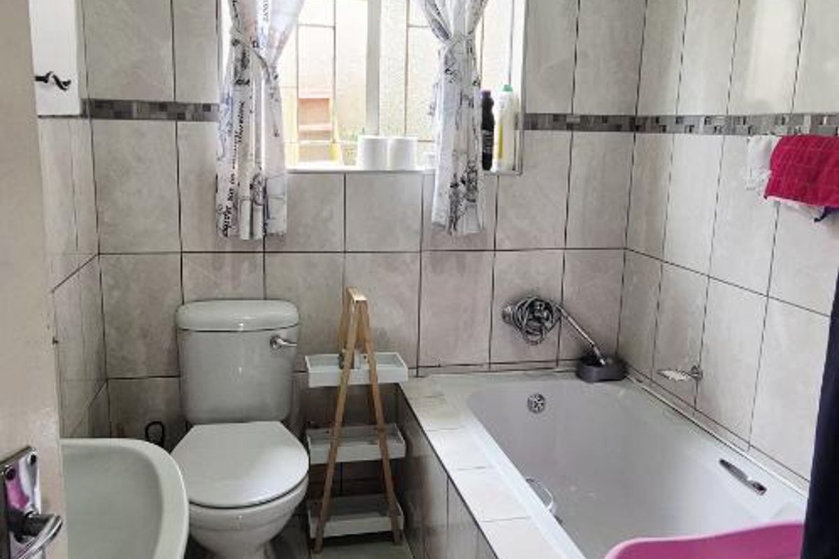 A functional bathroom featuring a built-in bathtub, ceramic wall and floor tiling, a standard toilet, and a window with security bars.