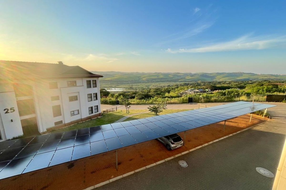 Elevated view of the apartment complex exterior, featuring a large solar panel carport structure, paved driveway, and scenic rolling hill landscape.
