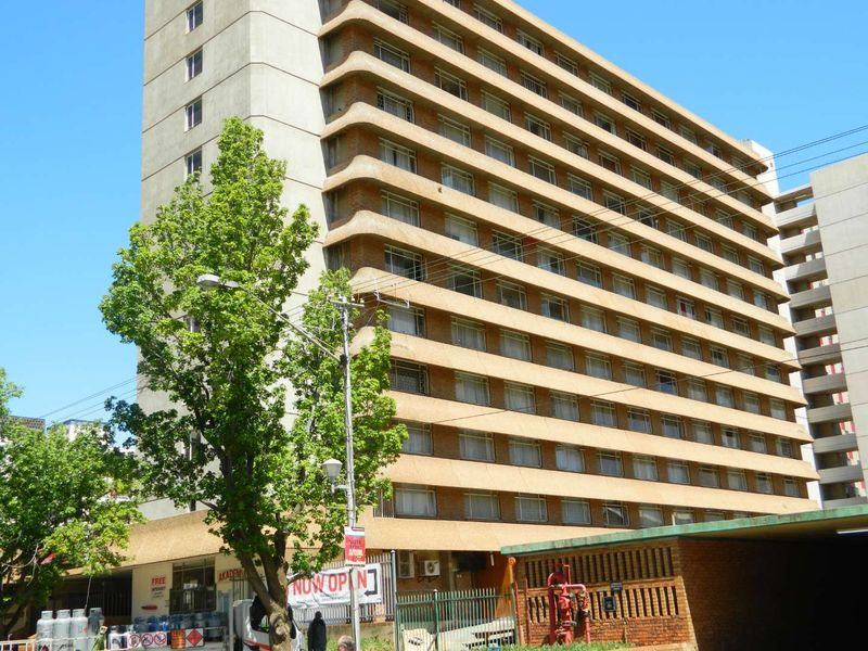 A multi-story apartment building exterior featuring a concrete facade with rows of balconies and ground-level security fencing.