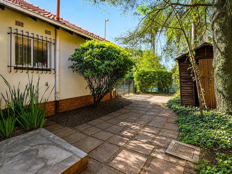 A rear exterior view featuring a paved walkway, established garden area, and secure window bars on the side of the house.