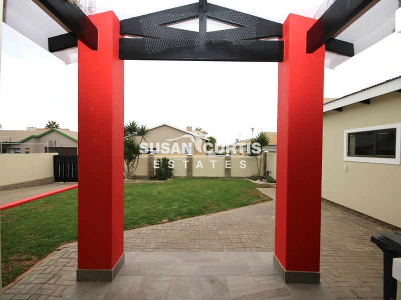 A covered patio area featuring a red-painted pergola structure, overlooking a private garden with paved pathways and boundary walls.