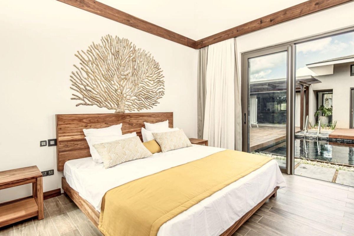A bright, modern bedroom featuring wood-look tile flooring, exposed timber ceiling beams, and large sliding glass doors opening directly onto a private pool deck.