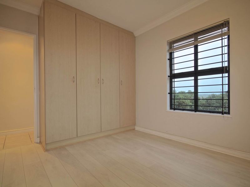 A bright, empty bedroom featuring light-toned laminate flooring, built-in cupboards, and a window fitted with security bars.