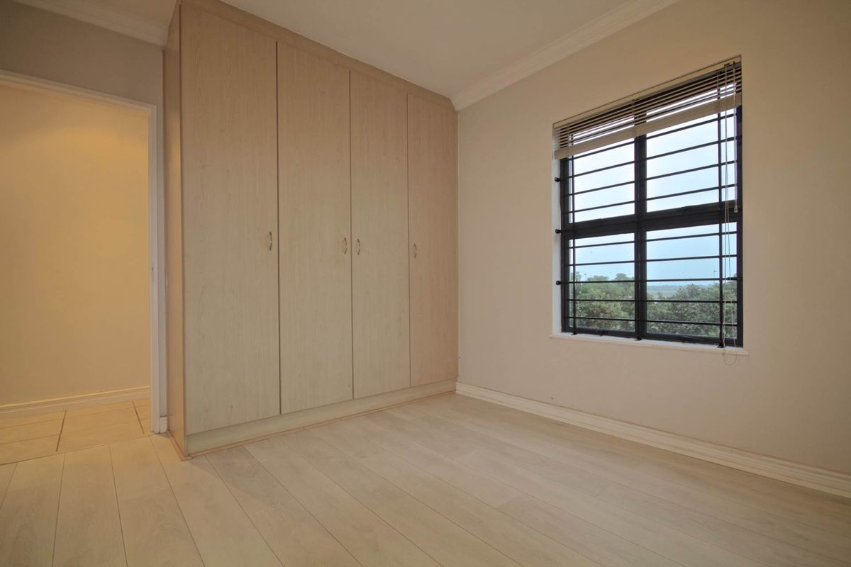 A bright, empty bedroom featuring light-toned laminate flooring, built-in cupboards, and a window fitted with security bars.