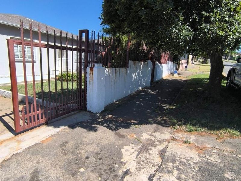 Front exterior view of the property featuring a metal security gate, perimeter wall, and paved driveway area showing signs of wear and rust.