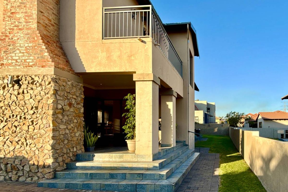 Rear elevation featuring a covered patio with tiled steps, stone-clad chimney, and an upper-level balcony with metal railings overlooking the garden.
