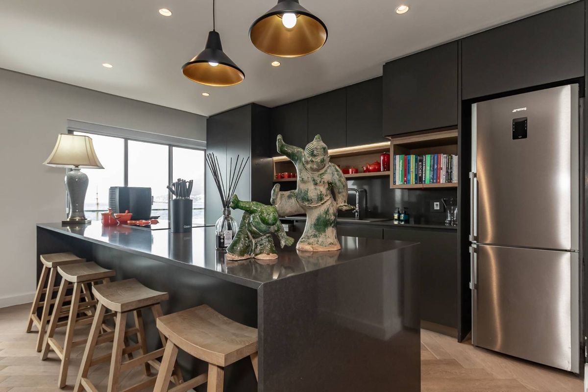 Modern open-plan kitchen featuring dark cabinetry, stone countertops, a breakfast island, integrated lighting, and stainless steel refrigerator.
