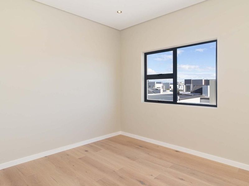 A bright, empty bedroom featuring modern laminate flooring, neutral walls, and a large aluminium-framed window with a neighbourhood view.