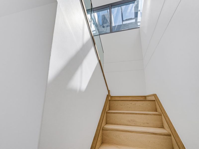 A bright, modern staircase featuring timber treads and a sleek glass balustrade, illuminated by a large overhead skylight.