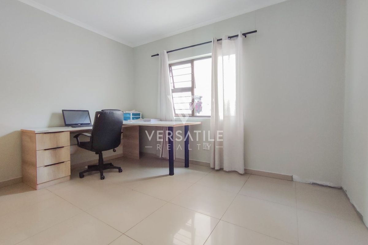A functional bedroom or home office space featuring neutral tiled flooring and a window with security bars.