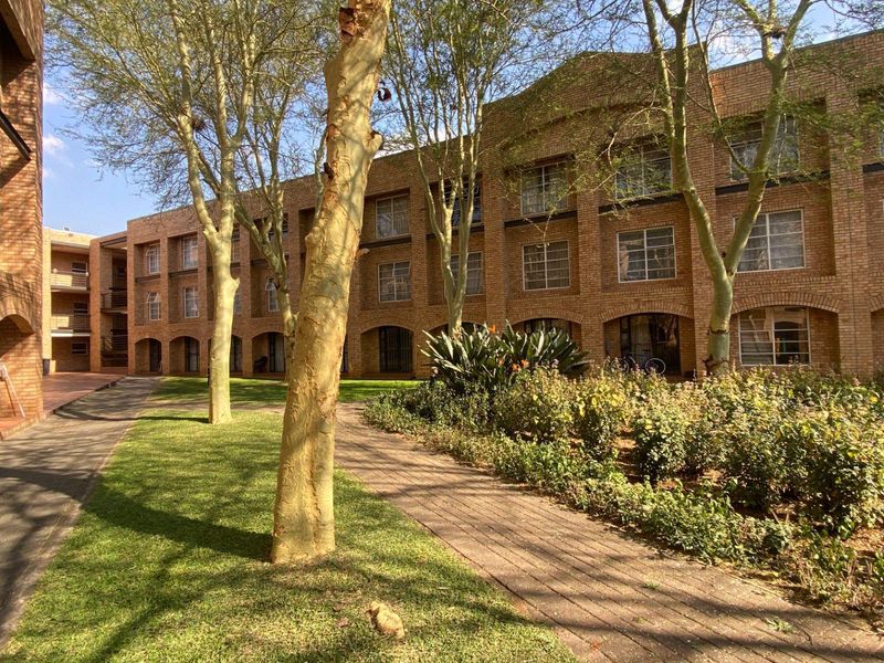 A view of the apartment complex exterior featuring face brick architecture, arched walkways, and well-maintained garden areas with mature trees.