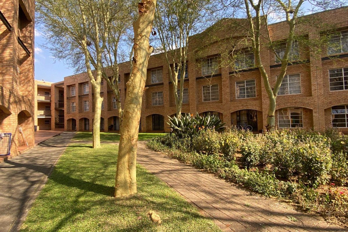 A view of the apartment complex exterior featuring face brick architecture, arched walkways, and well-maintained garden areas with mature trees.