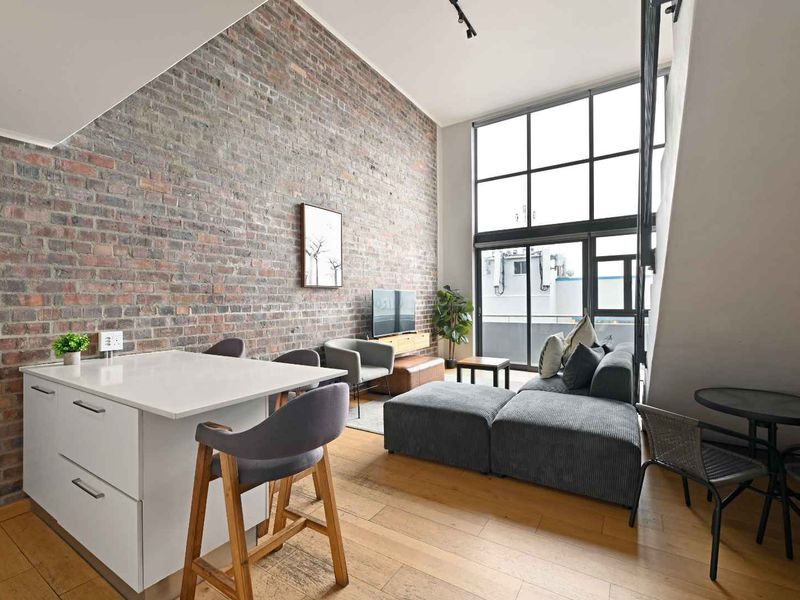 Modern open-plan living area featuring double-volume windows, exposed brick feature wall, and high-quality hardwood flooring in a contemporary penthouse setting.