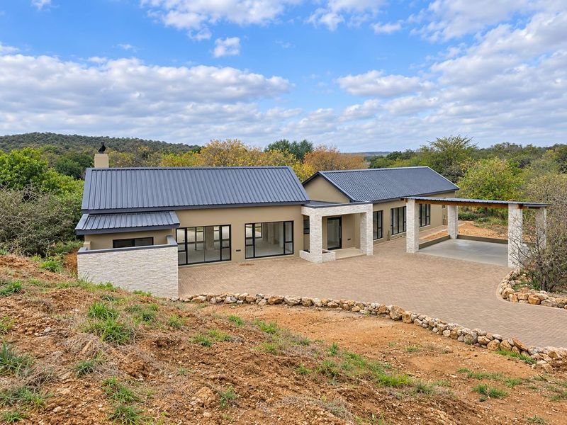 A modern single-story house featuring a metal roof, stone cladding accents, and a spacious paved driveway, set in a scenic natural landscape.