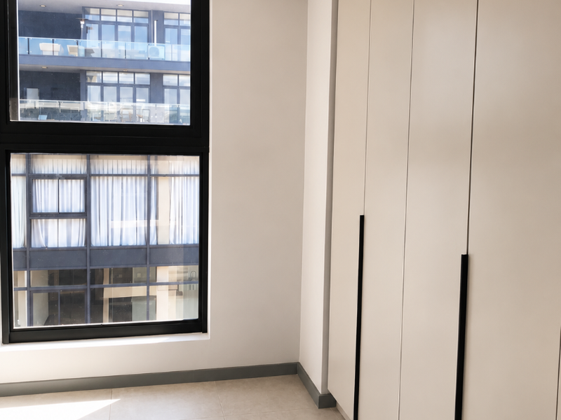 A bright, modern bedroom featuring neutral tiled flooring, floor-to-ceiling built-in cupboards with sleek black handles, and large aluminium-framed windows.