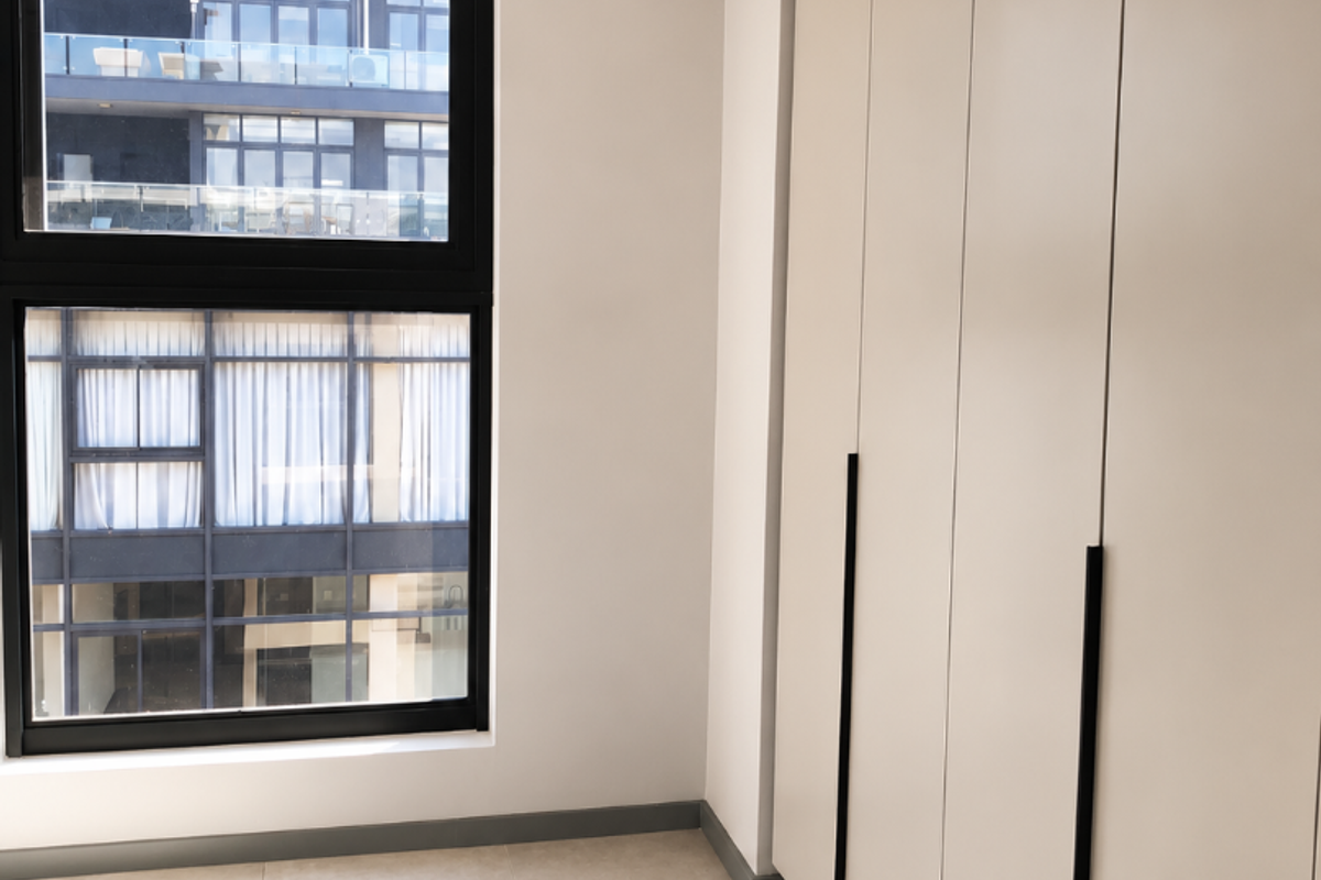 A bright, modern bedroom featuring neutral tiled flooring, floor-to-ceiling built-in cupboards with sleek black handles, and large aluminium-framed windows.