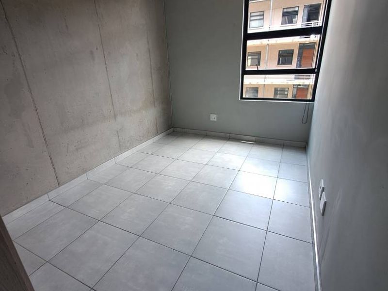 An empty bedroom featuring modern exposed concrete walls and ceiling with large windows and neutral tiled flooring.