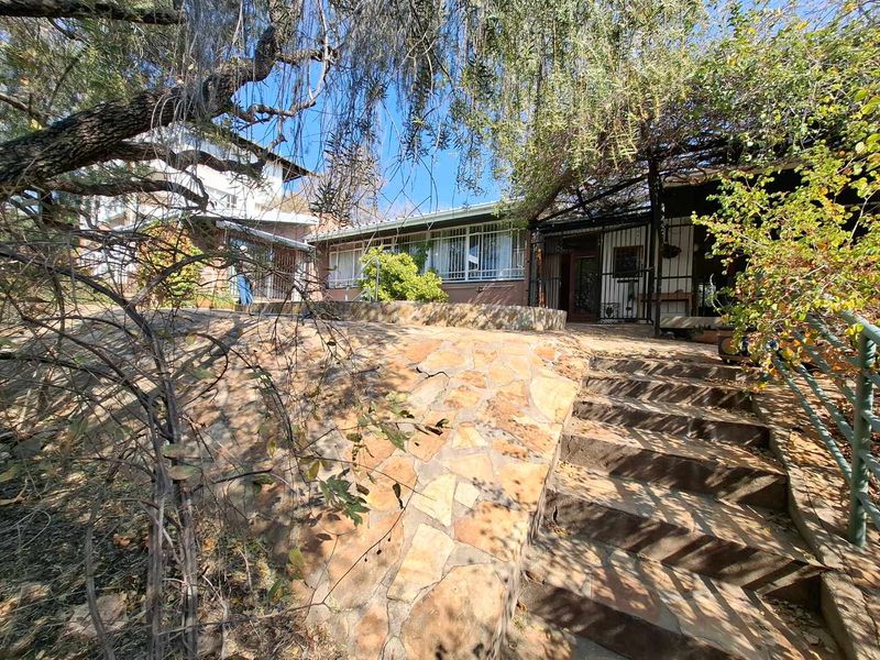 Rear exterior view featuring a stone retaining wall, stone staircase leading to a covered patio area, and mature garden landscaping.