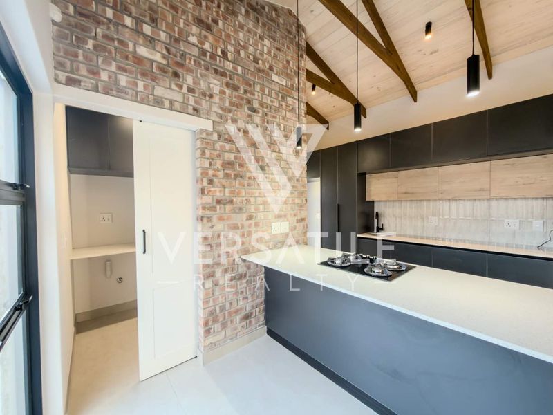 Modern kitchen featuring an exposed brick wall, timber ceiling beams, sleek cabinetry, stone countertops, and a convenient adjoining scullery area.