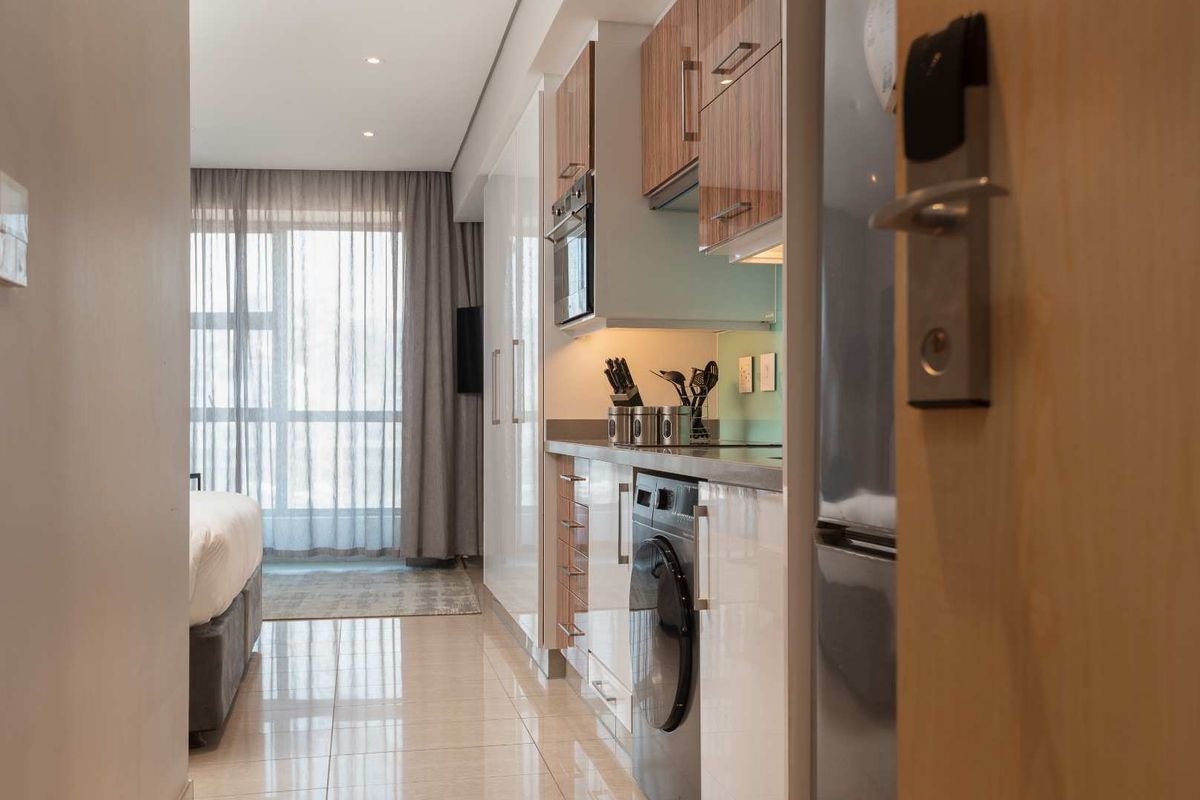 A compact, modern kitchen featuring high-gloss cabinetry, integrated appliances, and sleek tiled flooring, seamlessly flowing into the adjacent bedroom area.