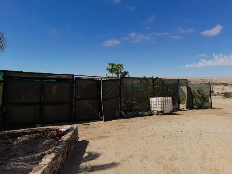 Outdoor area featuring shade netting structures, a water storage tank, and a stone retaining wall on a sandy plot.