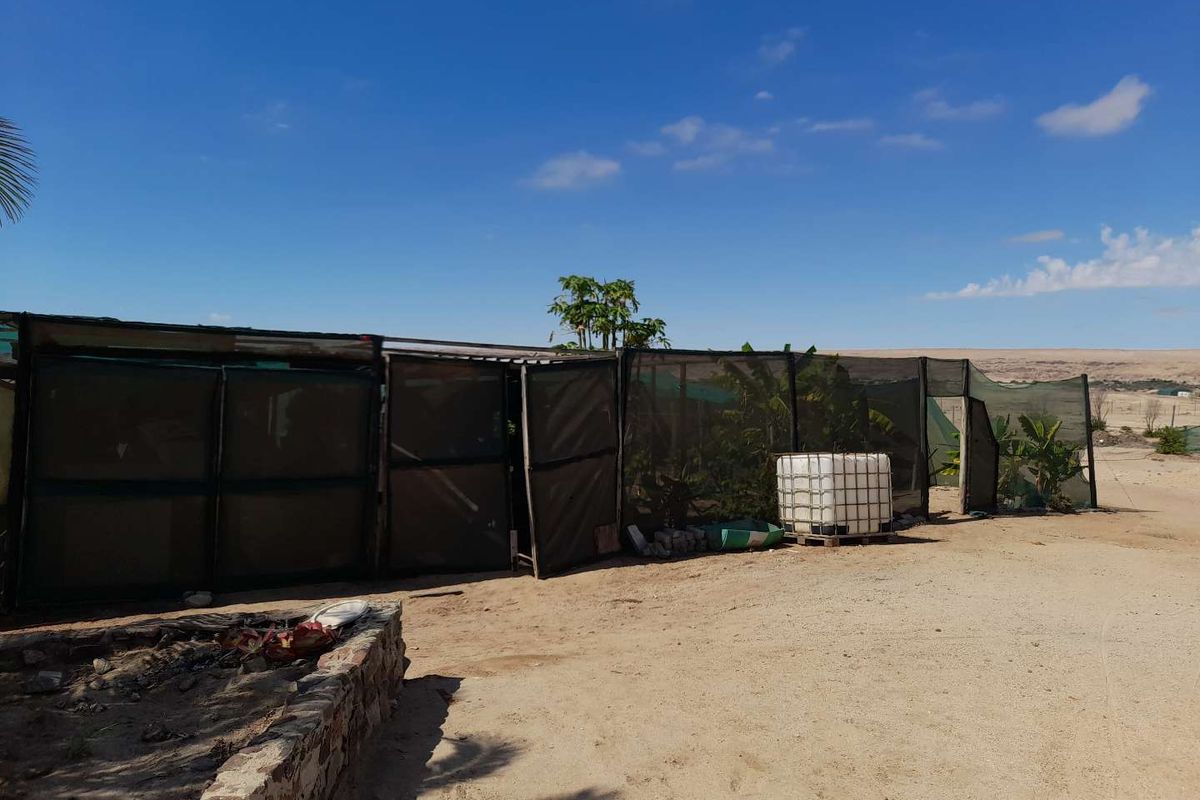 Outdoor area featuring shade netting structures, a water storage tank, and a stone retaining wall on a sandy plot.