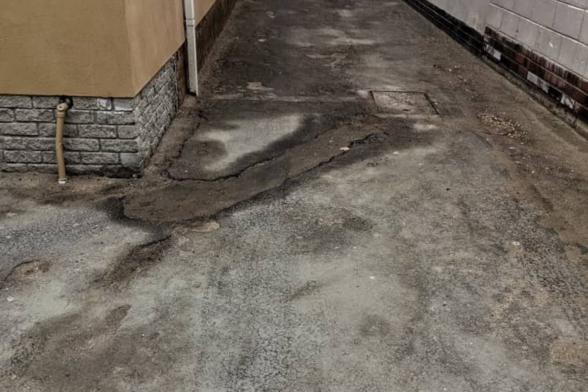 A narrow side passage or driveway area featuring concrete paving with visible cracks and signs of wear between the building and boundary wall.