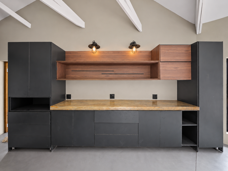 A modern kitchen area featuring dark matte cabinetry, a concrete countertop, and exposed white ceiling rafters, providing a clean and contemporary workspace.