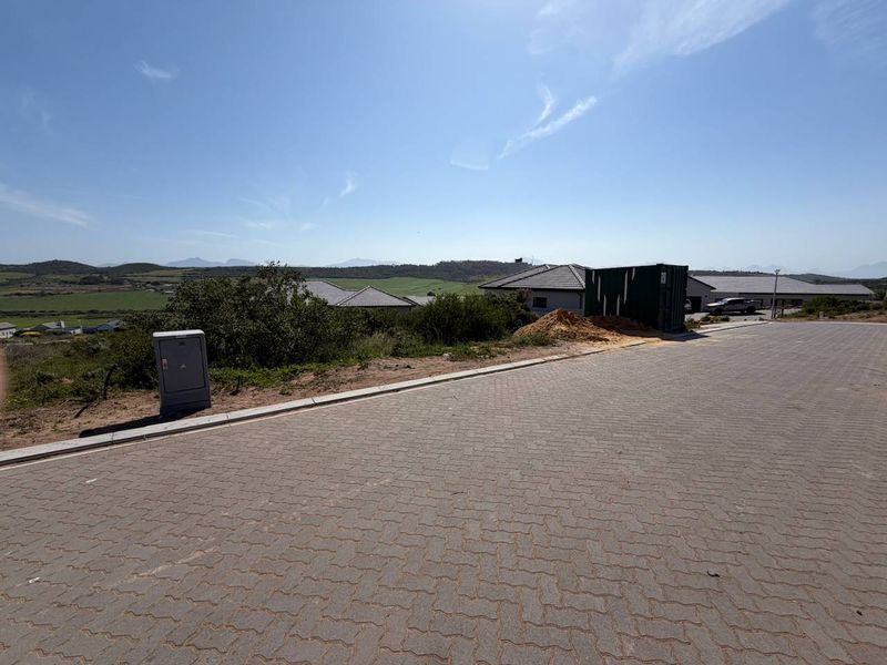 A street-level view of a residential vacant land plot featuring paved road access, kerbing, and an existing electrical utility connection point.