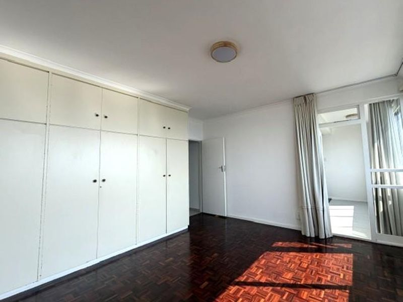 A spacious bedroom featuring classic parquet flooring, extensive built-in cupboards, and direct access to an adjoining balcony through a sliding glass door.