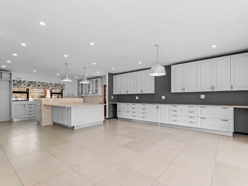 Spacious, modern kitchen featuring extensive white cabinetry, stone countertops, a central island, and neutral tiled flooring with recessed and pendant lighting.