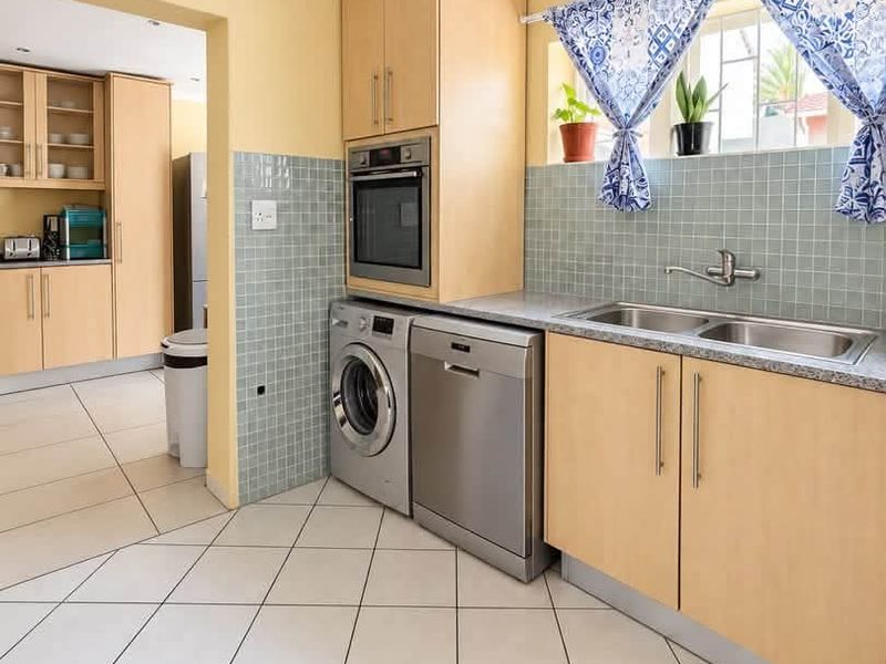 A functional scullery area featuring light wood cabinetry, tiled backsplash, double basin sink, and dedicated spaces for laundry and kitchen appliances.