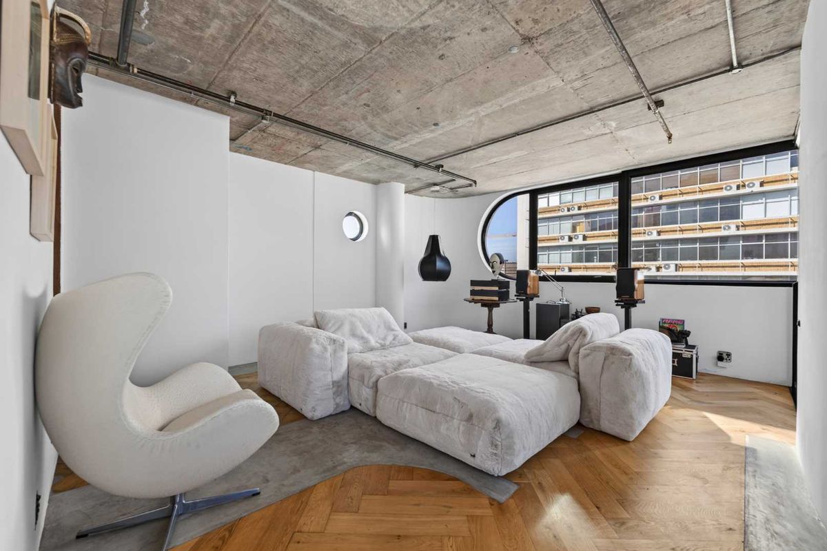 Modern living room featuring an exposed concrete ceiling, herringbone timber flooring, and a unique large feature window with an additional porthole window.