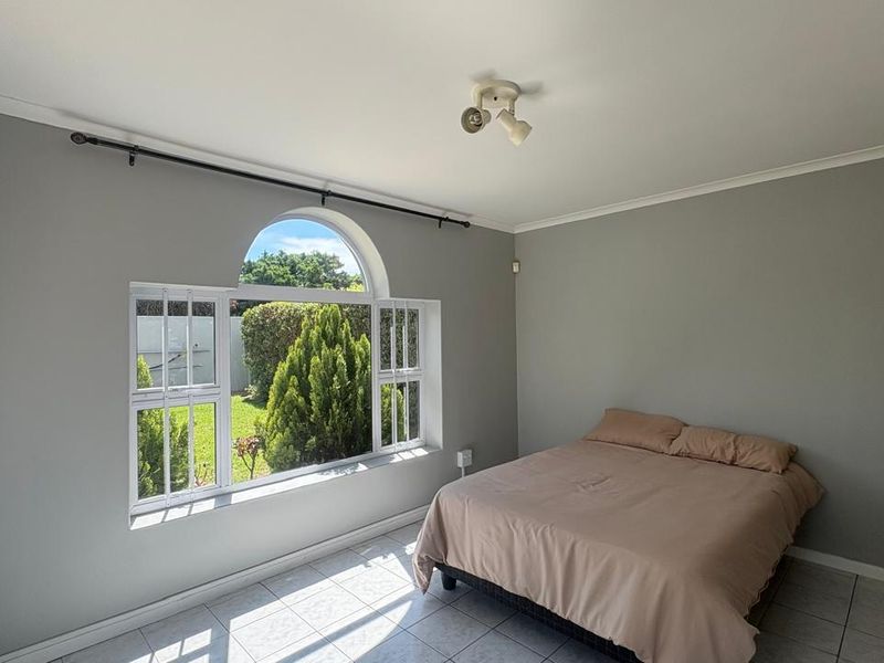 A bright bedroom featuring tiled flooring, a unique arched window with security bars, and neutral grey walls.