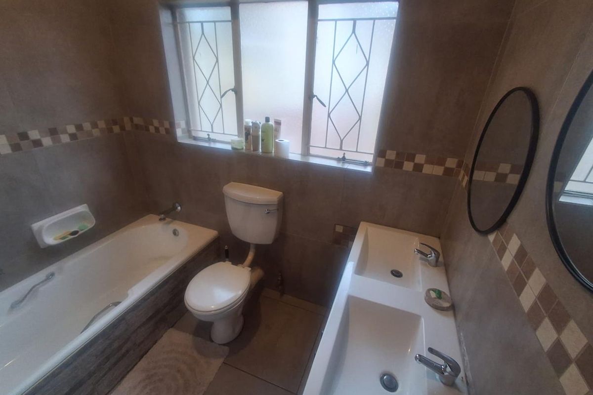 A functional bathroom featuring a bathtub, toilet, and a double vanity with tiled walls and natural light from a window with security bars.