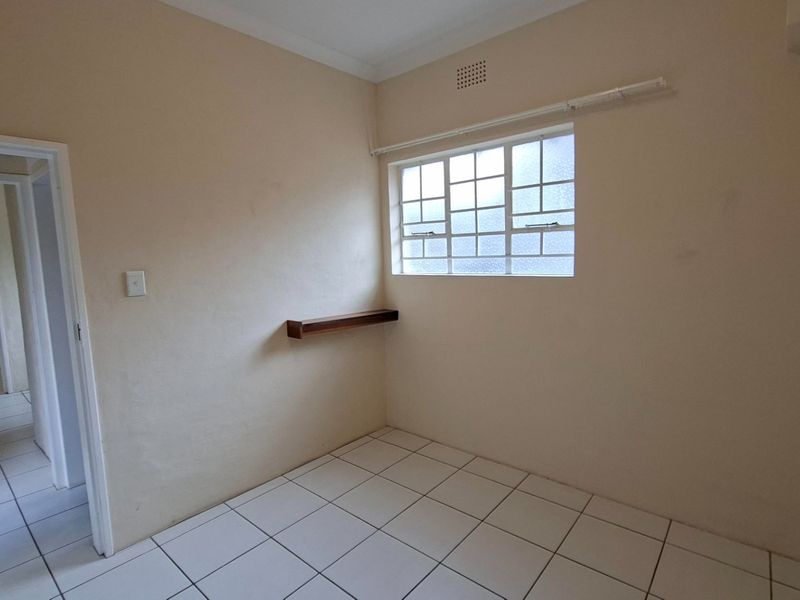 A clean, empty bedroom featuring white tiled flooring, a grid-pattern window, and a built-in wall shelf, offering a neutral space ready for personalization.