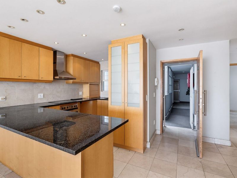 A functional kitchen featuring granite countertops, light wood cabinetry, integrated appliances, and tiled flooring, leading directly into the apartment hallway.