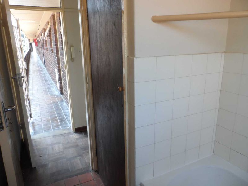 A dated bathroom interior featuring a bathtub with wall tiling, leading out to an external apartment corridor.
