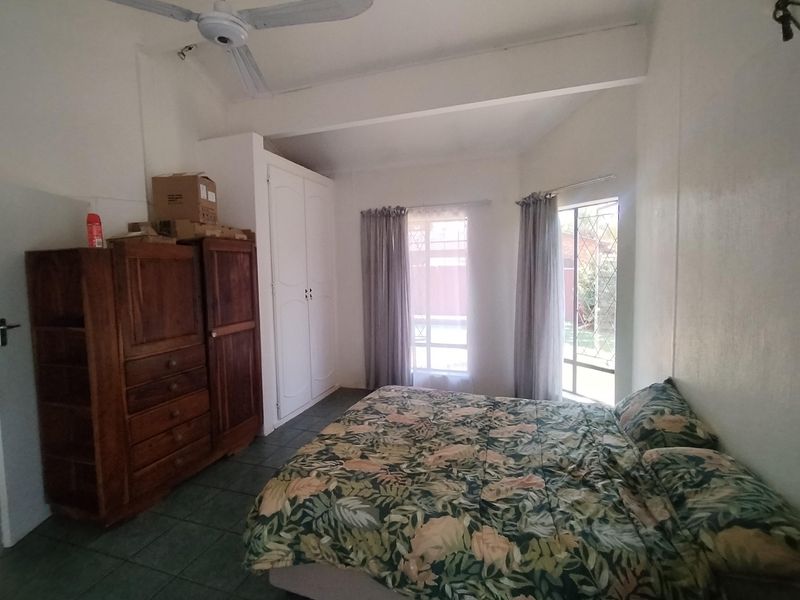 A functional bedroom featuring tiled flooring, a built-in wardrobe, a ceiling fan, and large windows with security bars allowing natural light.