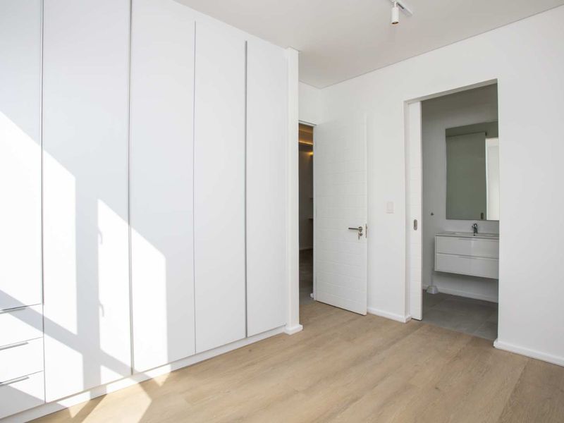 A bright, modern bedroom featuring extensive built-in cupboards, light laminate flooring, and direct access to an en-suite bathroom.