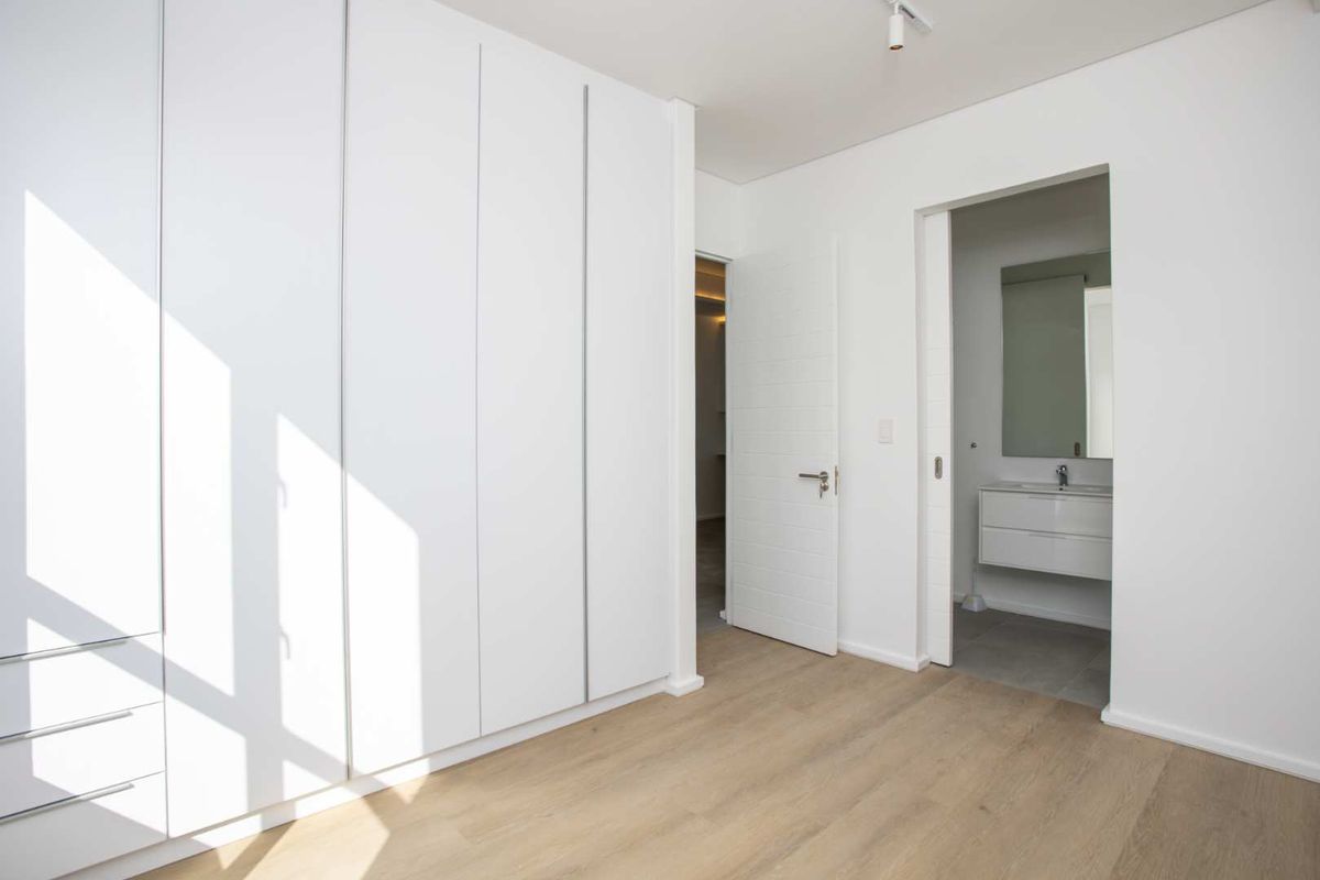 A bright, modern bedroom featuring extensive built-in cupboards, light laminate flooring, and direct access to an en-suite bathroom.
