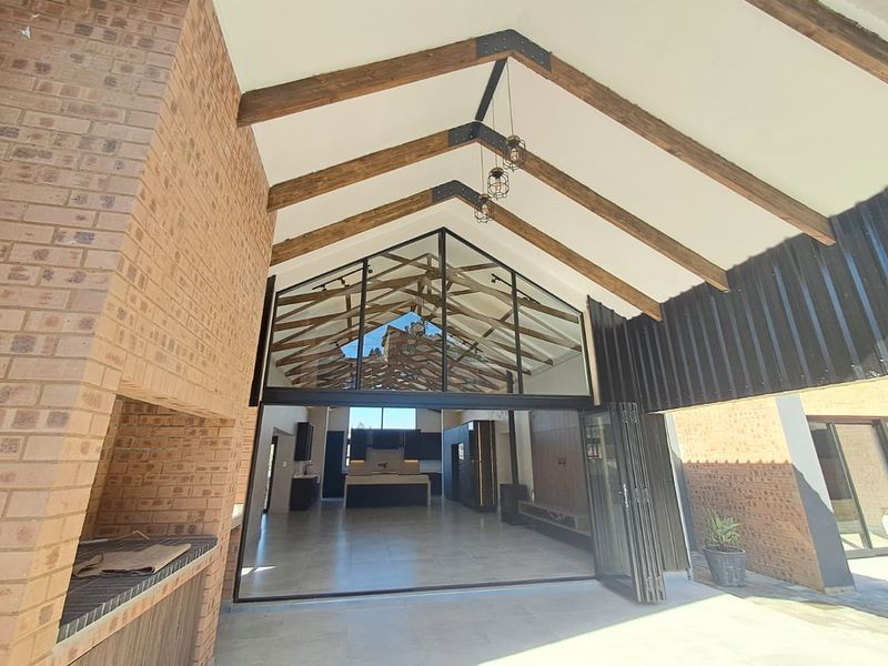 Spacious covered patio featuring exposed timber trusses, a built-in brick braai, and large stacking doors that open into the modern kitchen and living area.